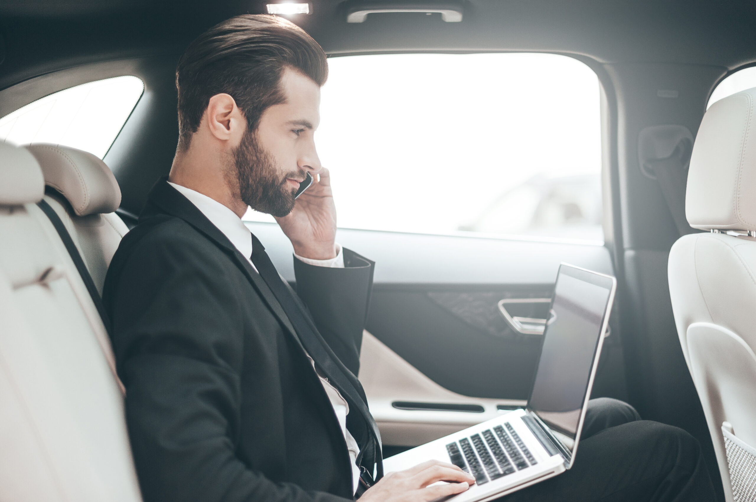 Confident young businessman working on his laptop and talking on the phone while sitting in the car